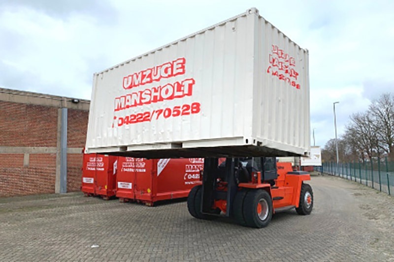 Ein roter Gabelstapler transportiert einen weißen Umzüge Mansholt Container an zwei roten Mansholt Containern vorbei