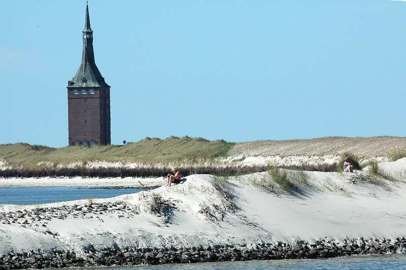 Ein sonnenbadender Mensch auf einer kleinen Sanddüne im Wattenmeer, im Hintergrund der Turm von Wangerooge