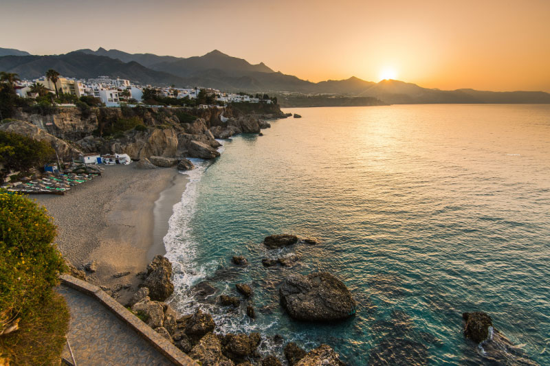Eine spanische Meeresbucht bei Sonnenuntergang mit einem Küstendorf im Hintergrund