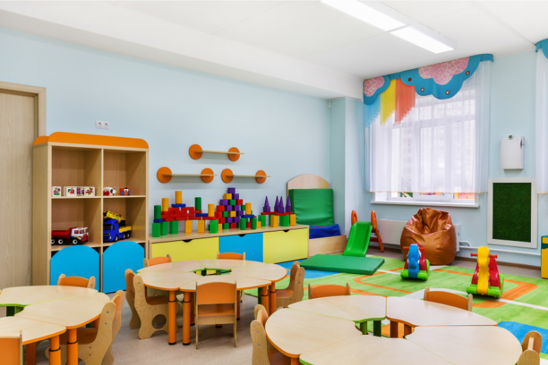 Das Foto zeigt das farbenfrohe Mobiliar eines Kindergartenraumes mit Holztischen, -stühlen und -schränken