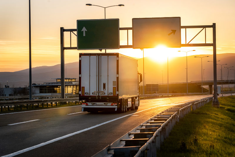 Ein LKW fährt auf der Autobahn in den Sonnenuntergang. Schilder weisen den Gütertransport entweder geradeaus oder nach rechts.