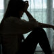In einem abgedunkelten Zimmer sitzt eine junge Frau auf dem Boden an die Couch gelehnt und stützt ihren Kopf in eine Hand