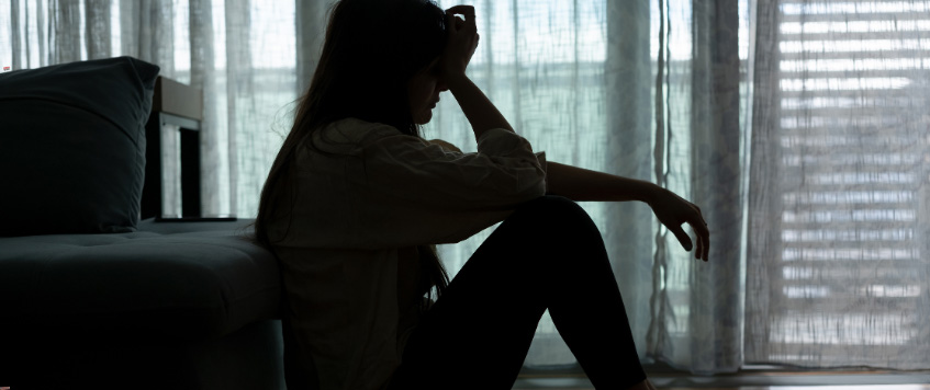 In einem abgedunkelten Zimmer sitzt eine junge Frau auf dem Boden an die Couch gelehnt und stützt ihren Kopf in eine Hand