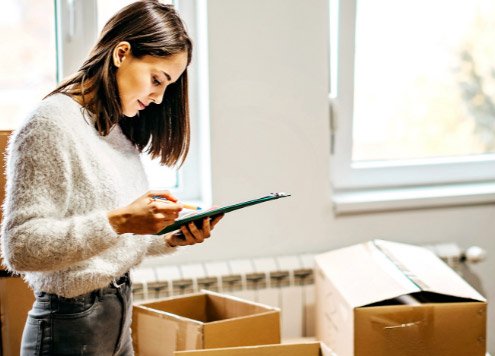 Eine Frau, die zwischen Umzugkartons steht, hält ein Klemmbrett in der Hand und hakt eine Checkliste darauf ab