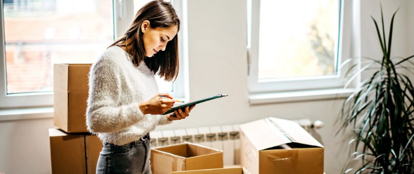 Eine Frau, die zwischen Umzugkartons steht, hält ein Klemmbrett in der Hand und hakt eine Checkliste darauf ab