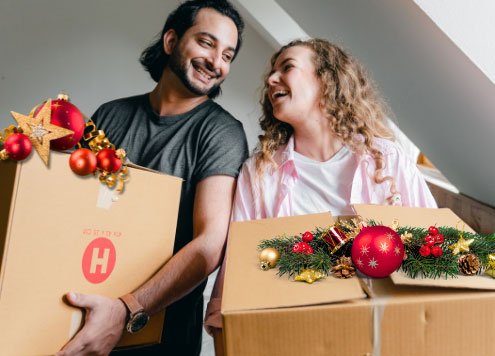 Ein Pärchen trägt jeweils einen Umzugskarton, der mit Weihnachtsschmuck dekoriert ist.