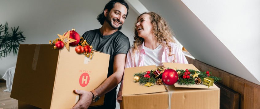 Ein Pärchen trägt jeweils einen Umzugskarton, der mit Weihnachtsschmuck dekoriert ist.