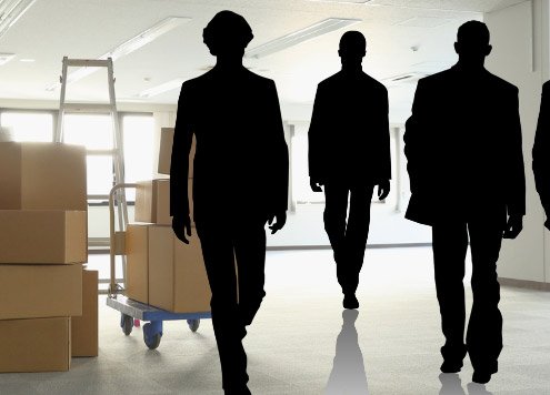 Mehrere schwarze Silhouetten gehen auf einen Stapel Umzugskartons in einem hellen Büro zu – Symbolbild für die Frage, wieviel Umzugshelfer man braucht