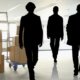 Mehrere schwarze Silhouetten gehen auf einen Stapel Umzugskartons in einem hellen Büro zu – Symbolbild für die Frage, wieviel Umzugshelfer man braucht