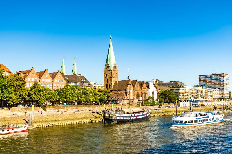 Blick auf die Weserpromenade in Bremen mit historischen Gebäuden, Kirche und Ausflugsschiffen bei sonnigem Wetter