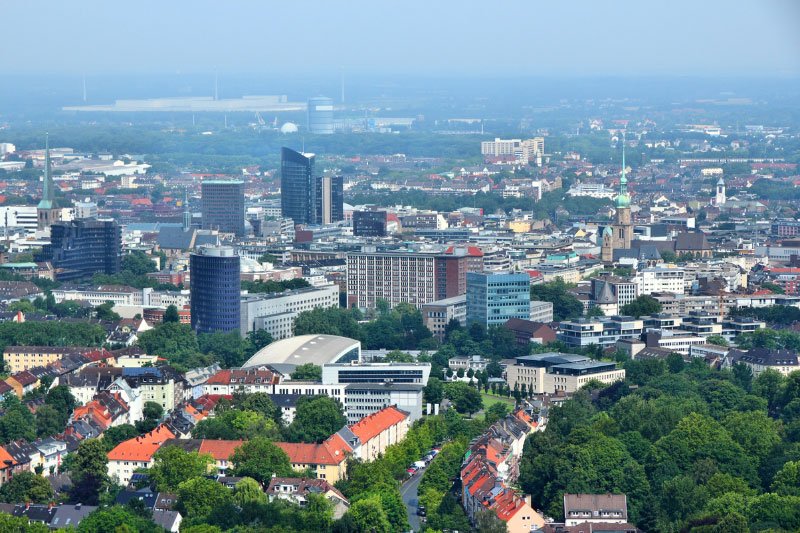 Panoramablick über die Stadt Dortmund mit moderner Innenstadtbebauung, Kirchen, Wohnvierteln und grünen Parkflächen bei klarem Himmel.