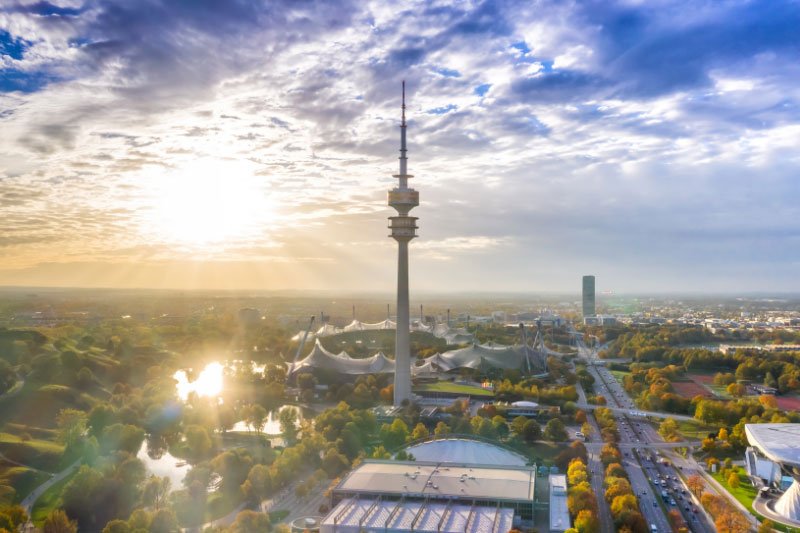 Blick über den Münchner Olympiapark mit Olympiaturm bei Sonnenaufgang und goldenem Licht über der Stadt.