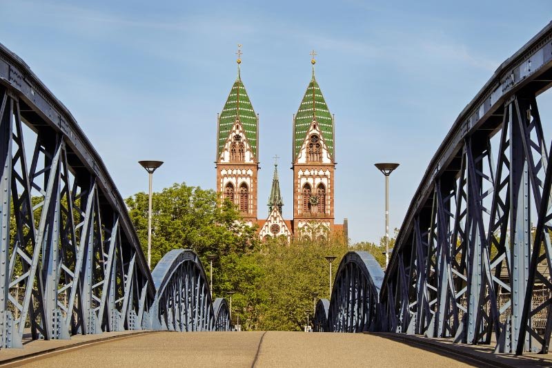 Freiburg-2 Blick über eine Brücke auf das Freiburger Münster mit seinen markanten Türmen, eingerahmt von Stahlkonstruktionen und Bäumen.