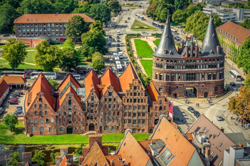 Blick auf das Holstentor in Lübeck mit den markanten Rundtürmen, davor historische Backsteinhäuser mit roten Giebeldächern und im Hintergrund Straßenverkehr sowie Grünanlagen