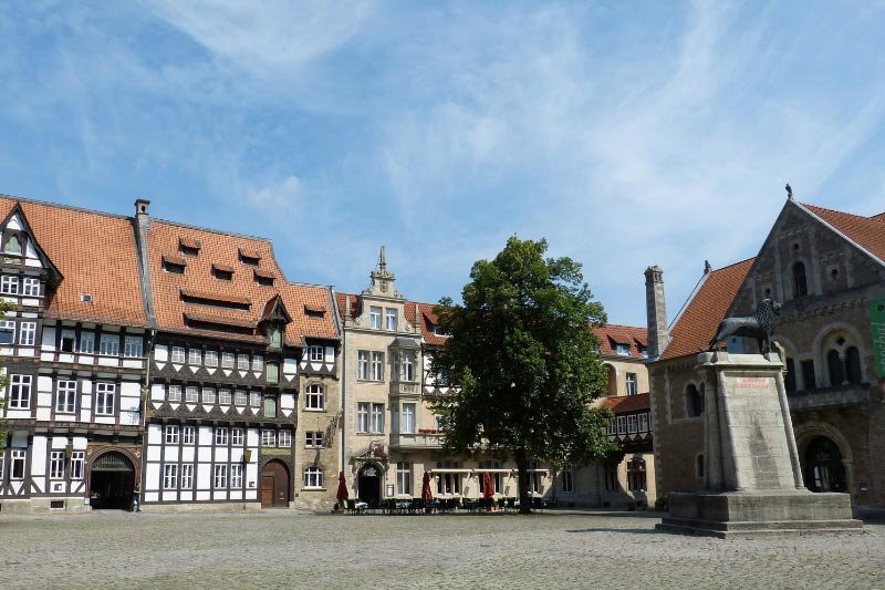 : Burgplatz in Braunschweig mit Fachwerkhäusern, Domfassade und Löwenstatue vor blauem Himmel.