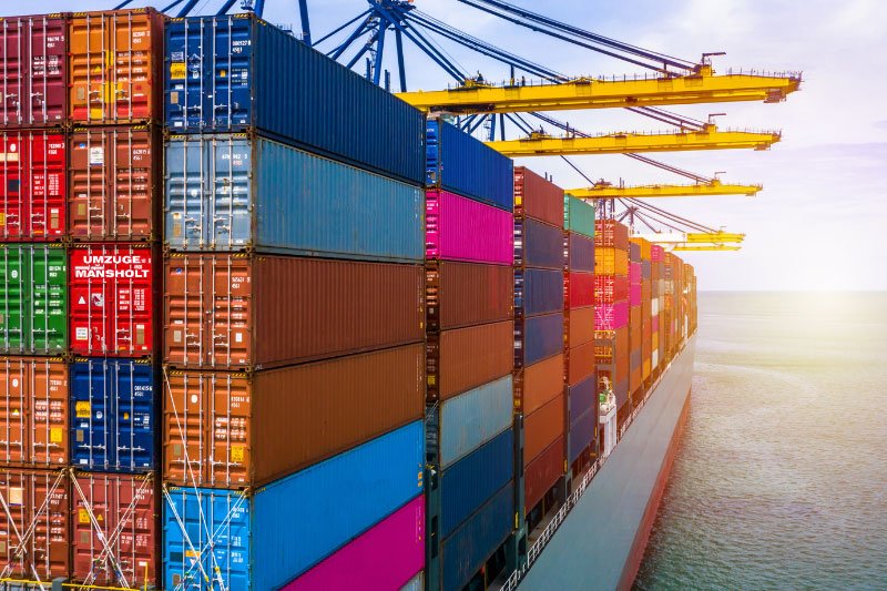 Gestapelte Container auf einem Frachtschiff im Hafen mit großen Verladekränen im Hintergrund, einer der Container trägt die Aufschrift Umzüge Mansholt