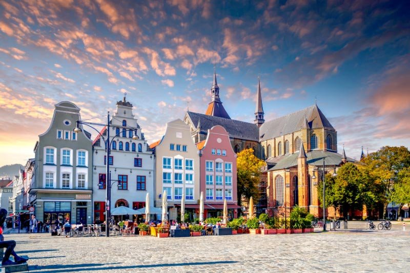 Historische Altstadthäuser und Kirche am Neuen Markt in Rostock bei sonnigem Himmel mit Cafés und belebtem Platz.