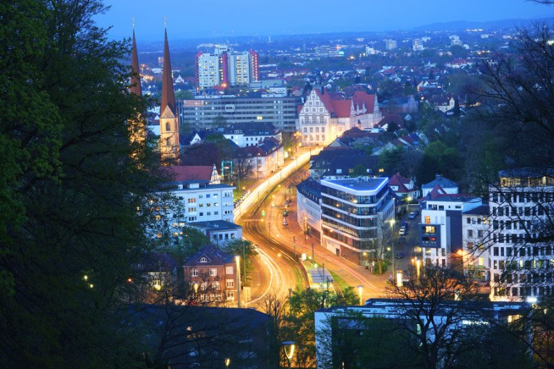 Abendlicher Blick über die Innenstadt von Bielefeld mit beleuchteten Straßen, Kirchtürmen und modernen Gebäuden vor der Kulisse des Teutoburger Waldes.]