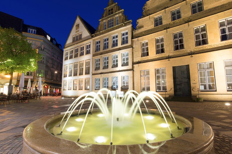 Beleuchteter Brunnen auf dem Alten Markt in Bielefeld vor historischen Bürgerhäusern und Restaurants in der Abenddämmerung