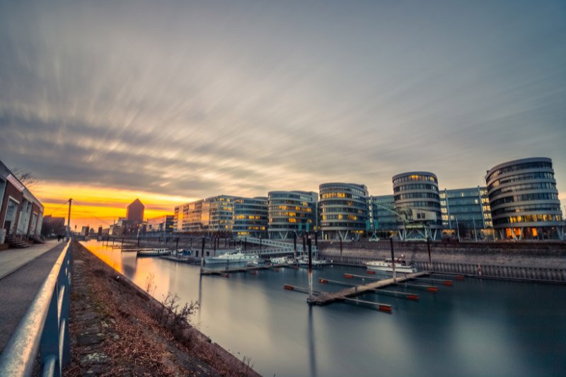Moderne Bürogebäude am Duisburger Innenhafen bei Sonnenuntergang mit ruhiger Wasserfläche, Anlegern und beleuchteten Fassaden unter dramatischem Abendhimmel