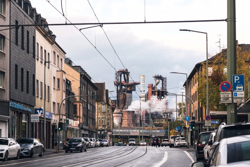 Straßenzug in Duisburg mit Straßenbahngleisen, Geschäften und Wohnhäusern, im Hintergrund die Industrieanlagen eines Stahlwerks mit aufsteigendem Dampf