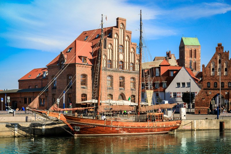 Historisches Segelschiff im Alten Hafen von Wismar vor den Backsteingebäuden der Altstadt und der St.-Marien-Kirche an einem sonnigen Tag.