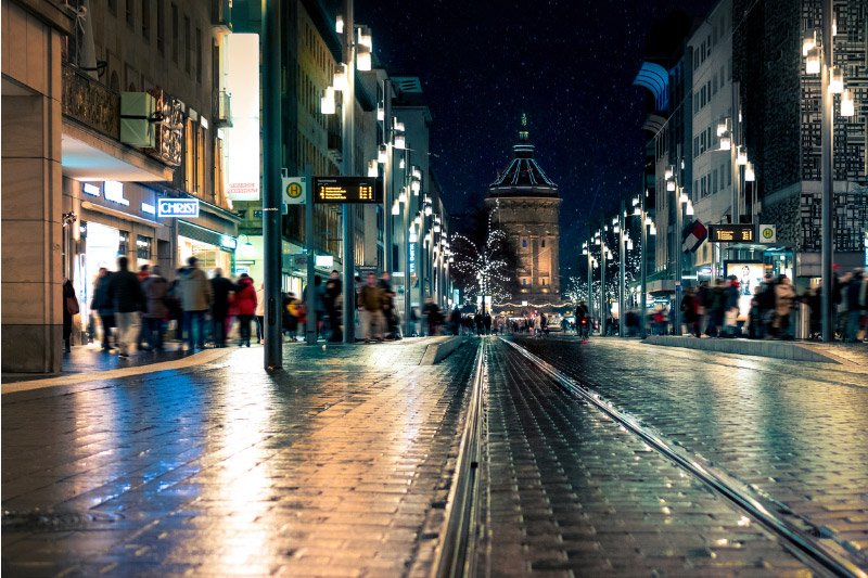 Belebte Einkaufsstraße in Mannheim bei Nacht mit Straßenbahngleisen und Blick auf den beleuchteten Wasserturm